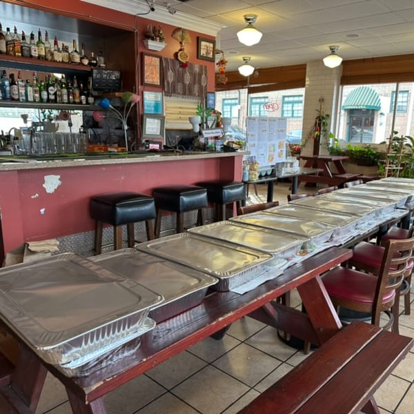 Long table filled with foil catering trays in a Thai restaurant dining area with a bar.