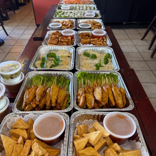 A long table filled with multiple foil trays of Thai appetizers, including spring rolls, satay, and fried tofu.