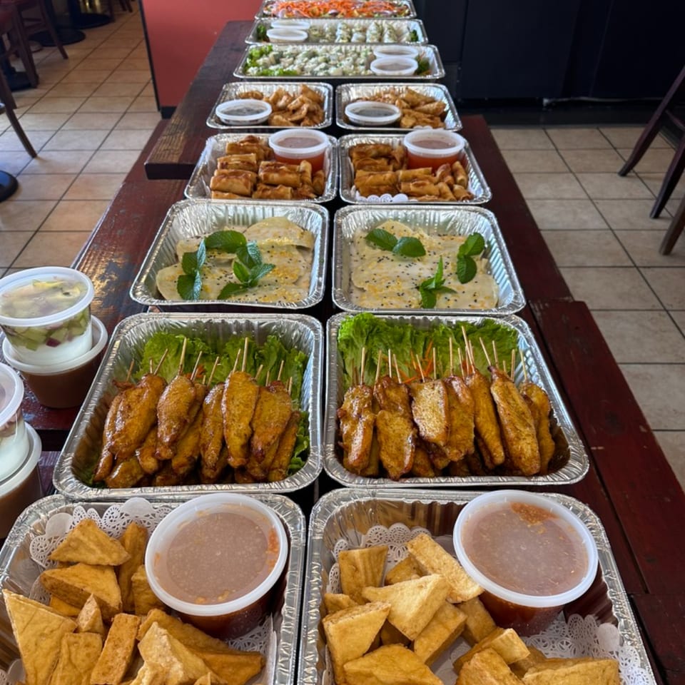 A long table filled with multiple foil trays of Thai appetizers, including spring rolls, satay, and fried tofu.