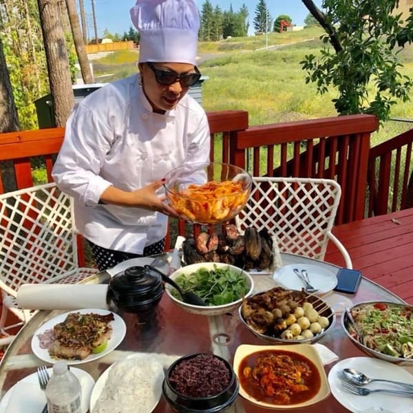 A chef in sunglasses and a white uniform holds a bowl of noodles over a table of Thai dishes.