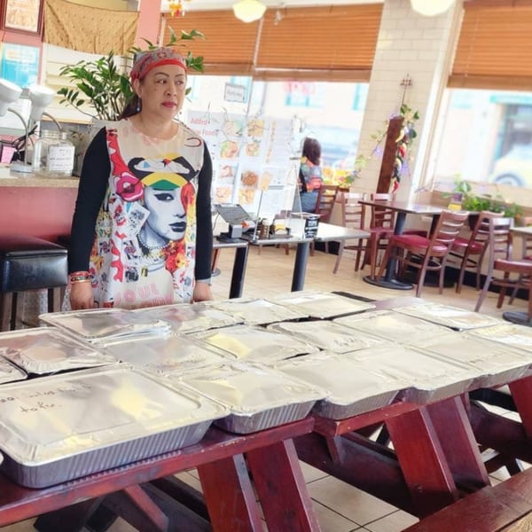 A woman in a colorful apron stands behind a table filled with foil takeout containers at Thai Chili Jam.