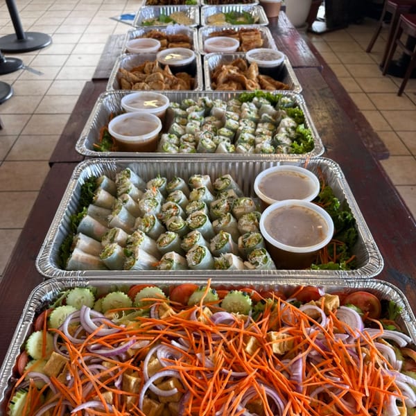 Multiple catering trays of Thai spring rolls, fried appetizers, and a fresh salad with shredded carrots.
