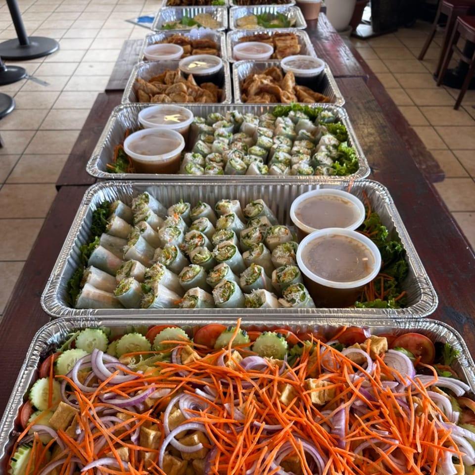 Multiple catering trays of Thai spring rolls, fried appetizers, and a fresh salad with shredded carrots.
